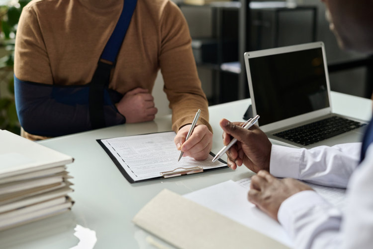 Amex Platinum Versicherungen - Mann mit Arm in einer Schlinge unterschreibt ein Formular, Doktor sitzt auf anderer Tischseite